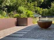 eine Feuerkugel aus rostigem Stahl. Sie steht auf einem steinigen Gartenplatz, rundherum ist viel grün.