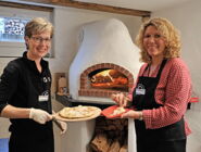 Zwei Frauen vor dem Pizzaofen, in der einen Hand einen fertig gebackenen Flammenkuchen.