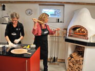 Eine Frau knetet den Pizzateig, eine zweite Frau schiebt eine belegte Pizza in den Ofen.